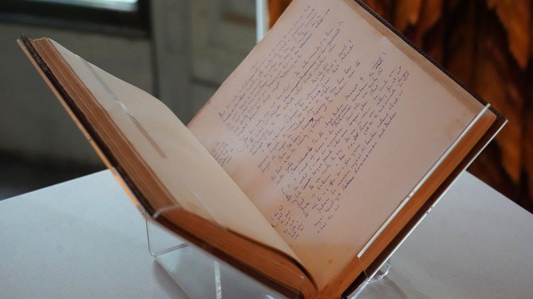 Photo of a book on a stand. The book is open and writing can be seen on the page.